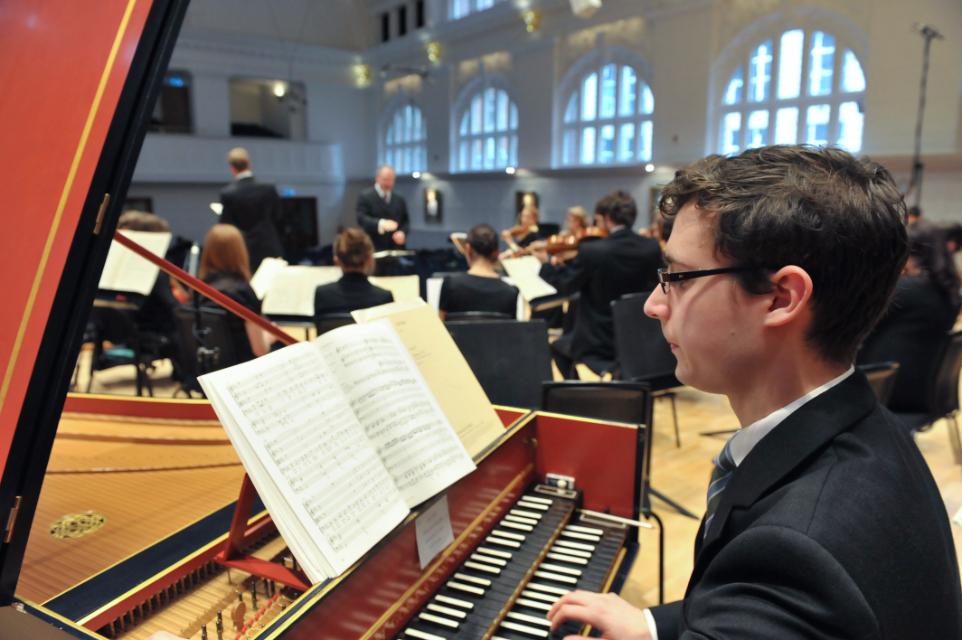 A male student with glasses, looking at the music sheets, playing the harpsichord, with an orchestra in the background, in a well lit background.